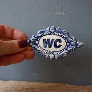 May include: A blue and white ceramic sign with the letters WC in a white oval. The sign is decorated with a blue and white floral design.