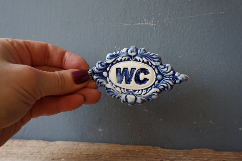 May include: A blue and white ceramic sign with the letters WC in a white oval. The sign is decorated with a blue and white floral design.