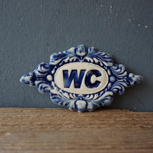 May include: A blue and white ceramic sign with a decorative border and the letters "WC" in the center.