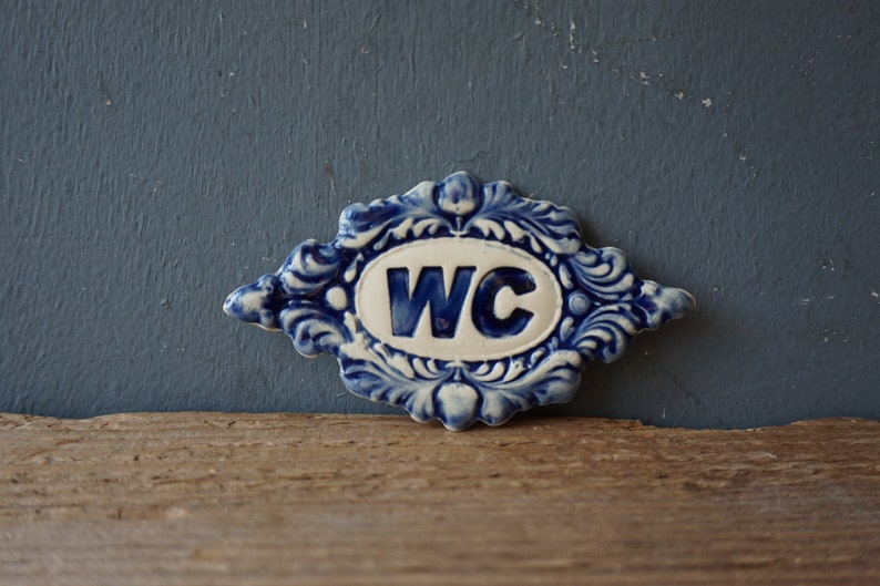 May include: A blue and white ceramic sign with a decorative border and the letters "WC" in the center.