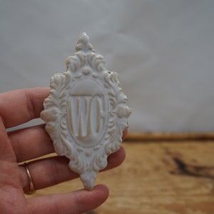 May include: White ceramic sign with an ornate frame and the letters "WC" in the center.