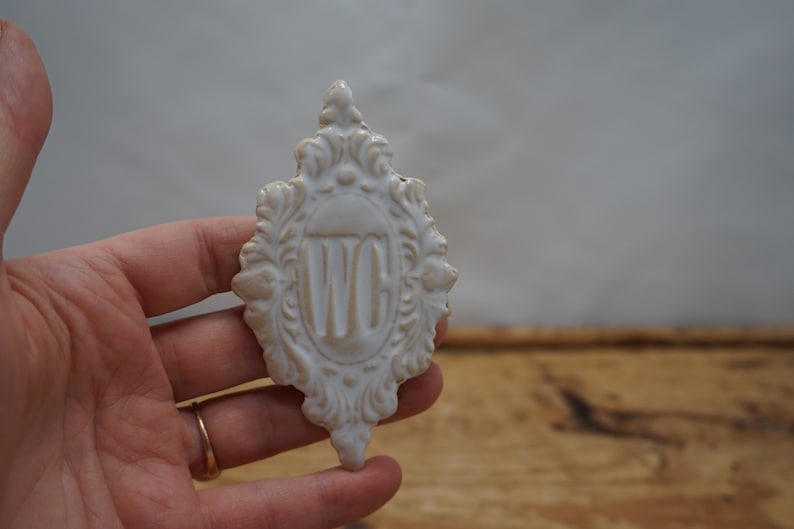 May include: White ceramic sign with an ornate frame and the letters "WC" in the center.