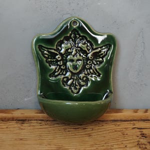 Könnte beinhalten: Eine grüne Keramik-Wandtasche mit einem dekorativen Engelsgesicht und Flügeldesign. Die Wandtasche hat ein kleines, gebogenes Becken am Boden und ein Loch oben zum Aufhängen. Das Stück ist glasiert und hat eine glänzende Oberfläche.