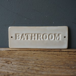 May include: Cream-colored rectangular bathroom sign with the word "BATHROOM" embossed in capital letters. The sign has two small holes and is set against a dark gray background and a wooden surface.