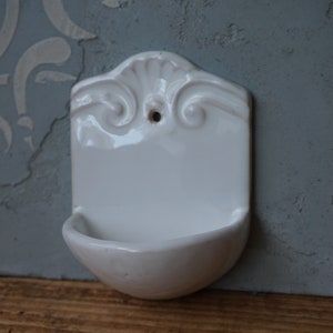 Pode incluir: Saboneteira de parede de cerâmica branca com uma borda decorativa festoneada e um pequeno furo na parte de trás para pendurar.