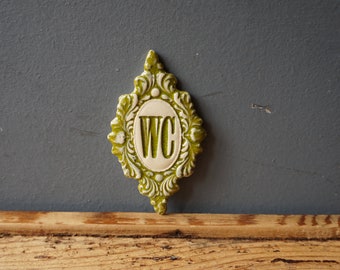 May include: A green and white ceramic sign with a decorative frame and the letters "WC" in the center.
