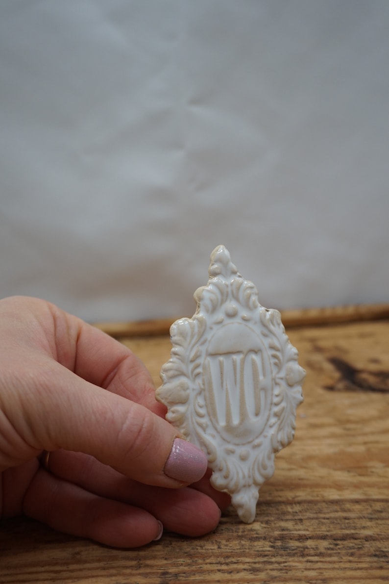 May include: White ceramic sign with an ornate frame and the letters "WC" in the center.