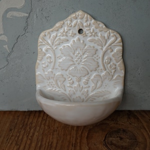 May include: White ceramic wall mounted soap dish with a floral design. The dish has a scalloped edge and a small hole at the top for hanging.