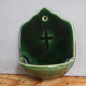 Może przedstawiać: Ceramiczna kropielnica ścienna z błyszczącą glazurą w kolorze głębokiej leśnej zieleni. Przedmiot ma kształt tarczy z małym okrągłym otworem u góry do zawieszenia oraz wypukłym symbolem krzyża umieszczonym nad zaokrąglonym zbiornikiem na wodę.