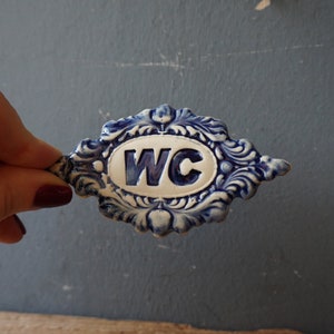May include: A blue and white ceramic sign with a decorative border and the letters "WC" in the center.