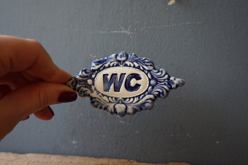 May include: A blue and white ceramic sign with a decorative border and the letters "WC" in the center.