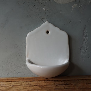May include: A white ceramic wall-mounted basin with a rounded bottom and a small hole at the top for hanging. The basin has a smooth, glossy finish and is set against a gray wall.