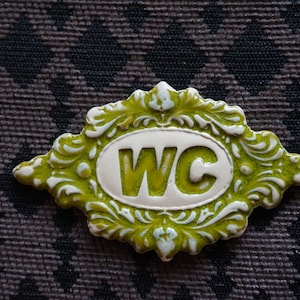 May include: A green and white ceramic sign with an ornate frame and the letters "WC" in the center.