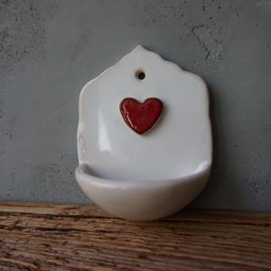 Op de afbeelding: Een witte keramische wandzak met een rood hart in het midden. Het hart is verhoogd en getextureerd. De zak heeft een kleine, gebogen kom aan de onderkant en een gat aan de bovenkant om op te hangen. De achtergrond is een grijze muur.