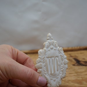 May include: White ceramic sign with an ornate frame and the letters "WC" in the center.