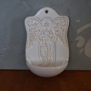 May include: A glossy off-white ceramic wall-mounted holy water font featuring a raised relief design of an angel with wings and hands pressed together in prayer. The piece has a rounded basin at the bottom and a small hole at the top for hanging.