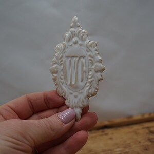May include: White ceramic sign with an ornate frame and the letters "WC" in the center. The sign is held in a hand.