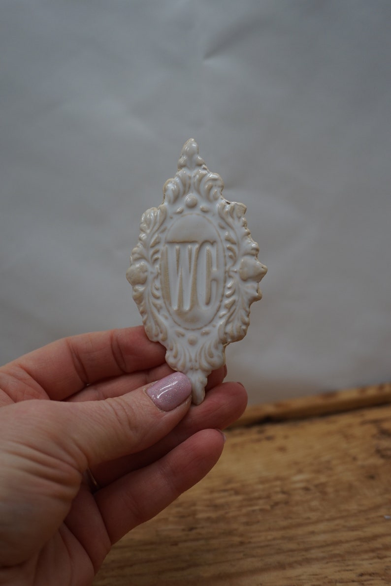 May include: White ceramic sign with an ornate frame and the letters "WC" in the center. The sign is held in a hand.