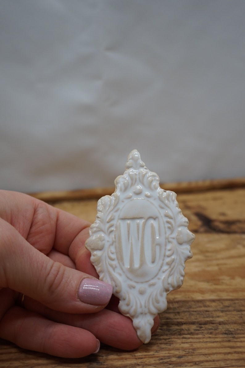May include: White ceramic sign with an ornate frame and the letters "WC" in the center. The sign is being held in a hand.