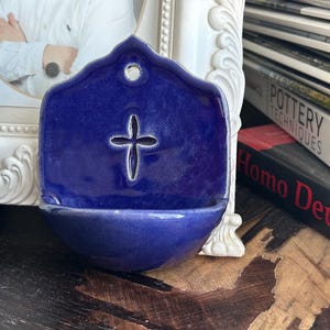May include: A cobalt blue ceramic holy water font with a cross cutout. The wall-mounted font has a rounded basin and a hole for hanging. The background includes a framed picture and books.