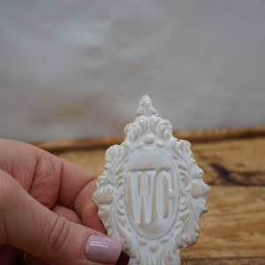 May include: White ceramic sign with an ornate frame and the letters "WC" in the center. The sign is being held in a hand.