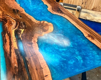 May include: A wooden table top with a blue epoxy river running through the center. The wood is a dark brown with lighter brown streaks. The epoxy is a bright blue with white swirls.