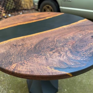May include: A round table featuring a natural wood grain surface with a prominent dark black resin inlay running across the center and edges. The table is supported by a solid black geometric pedestal base, showcasing a contrast between wood and resin.