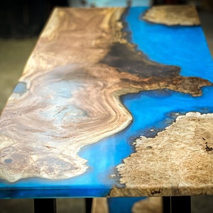 May include: A wooden table top with a blue epoxy resin inlay. The resin creates a swirling pattern that resembles a river or ocean. The wood is a light brown color with dark brown streaks.
