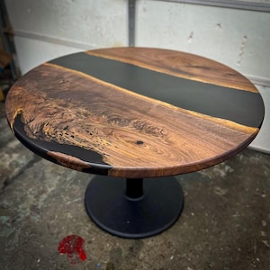 May include: A round table featuring a natural wood slab with a prominent live edge and a dark, opaque resin inlay running through the center. The tabletop is supported by a single, solid black pedestal base with a circular floor plate.
