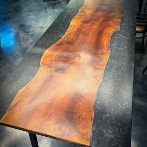 May include: A long, wooden table top with a natural wood grain finish. The table top is split in half, with one half being a dark gray epoxy resin. The table top is supported by a black metal base with wheels.