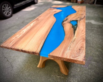 Pode incluir: Uma mesa de madeira com um rio de epóxi azul que atravessa o centro. A mesa tem um acabamento de madeira natural e é suportada por uma base de madeira.