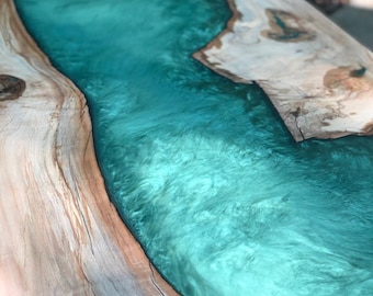 May include: A close-up of a wooden table top with a large, flowing river of turquoise epoxy resin. The resin has a shimmering, iridescent effect.