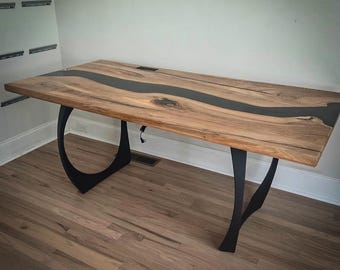 May include: A rectangular desk featuring a natural wood slab tabletop with a dark, flowing resin inlay running through the center. The desk is supported by two black, curved metal legs and includes a rectangular power outlet port on the surface.