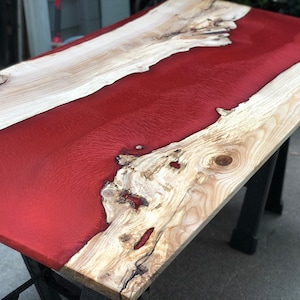 May include: A wooden table top with a red epoxy resin river running through the center. The wood is a light brown color and the resin is a deep red.