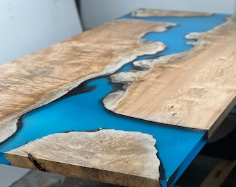 May include: A wooden table top with a blue epoxy river running through it. The wood has a natural grain pattern and the epoxy is smooth and glossy.