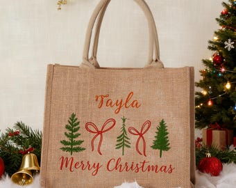 May include: A beige burlap tote bag with a Christmas theme. The bag has two handles and features the name "Tayla" in red script, along with green Christmas trees and red bows. The words "Merry Christmas" are also in red.