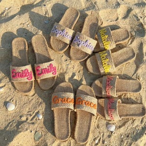 Puede incluir: Un conjunto de sandalias de yute personalizadas con nombres bordados en varios colores. Las sandalias están dispuestas sobre arena, mostrando los nombres "Sophia", "Emily", "Natalie", "Madison" y "Grace". Las sandalias son de color beige natural.
