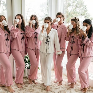Könnte beinhalten: Eine Gruppe von Personen in passenden rosa Pyjamas mit schwarzen Verzierungen und Schleifen, die weiße Tassen halten. Eine Person trägt einen weißen Pyjama mit schwarzen Verzierungen. Die Pyjamas haben lange Ärmel und Hosen. Die Szene spielt sich in einem Innenraum ab.