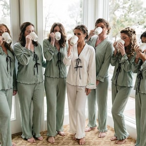 Pode incluir: Sete pessoas vestindo pijamas combinando bebem de canecas brancas. Seis estão em pijamas verde claro com acabamentos e laços pretos, enquanto uma pessoa usa pijamas brancos com acabamentos e laços pretos. Os pijamas têm bainha com babados.