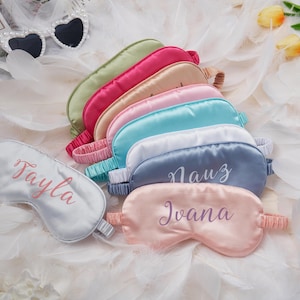 Könnte beinhalten: Eine Auswahl an Satin-Schlafmasken in verschiedenen Farben, darunter Rosa, Blau und Grün, mit gestickten Namen. Die Masken haben elastische Bänder und liegen auf weißen Federn. Herzförmige Sonnenbrillen sind im Hintergrund zu sehen.