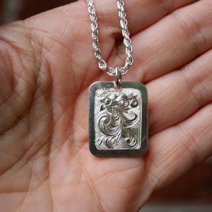 Puede incluir: Un colgante rectangular de tono plateado que cuelga de una cadena de estilo cuerda trenzada. La cara del colgante presenta un intrincado diseño floral y de volutas en relieve, con un acabado metálico pulido y grabados detallados.