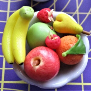 May include: A small white bowl filled with miniature, colorful fruit. The arrangement includes yellow bananas, a green apple, a red apple, a strawberry, a pear, and an orange with a green leaf. The fruit is arranged on a purple and yellow checkered surface.