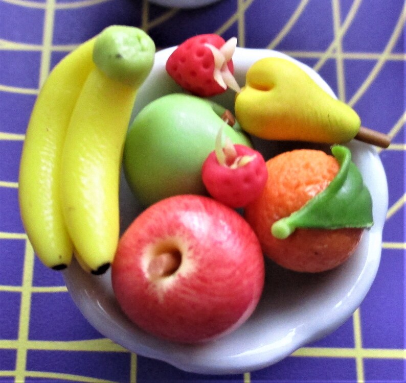 May include: A small white bowl filled with miniature, colorful fruit. The arrangement includes yellow bananas, a green apple, a red apple, a strawberry, a pear, and an orange with a green leaf. The fruit is arranged on a purple and yellow checkered surface.
