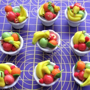 May include: Nine miniature white bowls filled with colorful, handcrafted fruit. Each bowl contains a variety of fruits, including bananas, apples, oranges, pears, and strawberries. The fruits are in shades of red, green, yellow, and orange. The bowls are arranged on a purple surface.