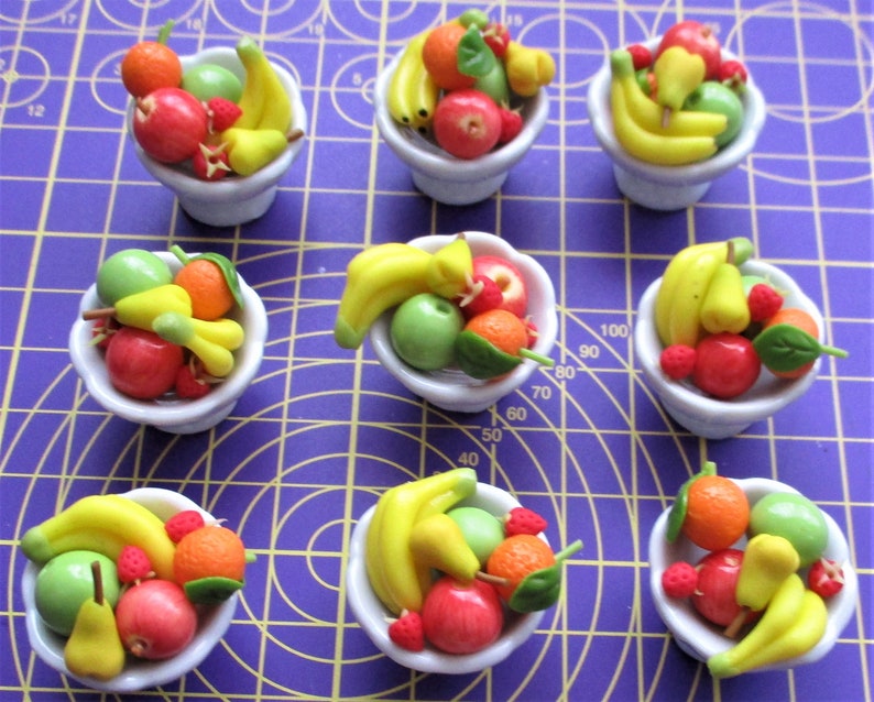 May include: Nine miniature white bowls filled with colorful, handcrafted fruit. Each bowl contains a variety of fruits, including bananas, apples, oranges, pears, and strawberries. The fruits are in shades of red, green, yellow, and orange. The bowls are arranged on a purple surface.