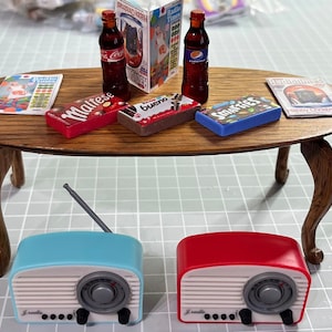 Peut inclure: Une table miniature avec deux radios vintage, une bleue et une rouge, avec une antenne argentée. La table est décorée de barres chocolatées miniatures, de magazines et de deux bouteilles de soda.