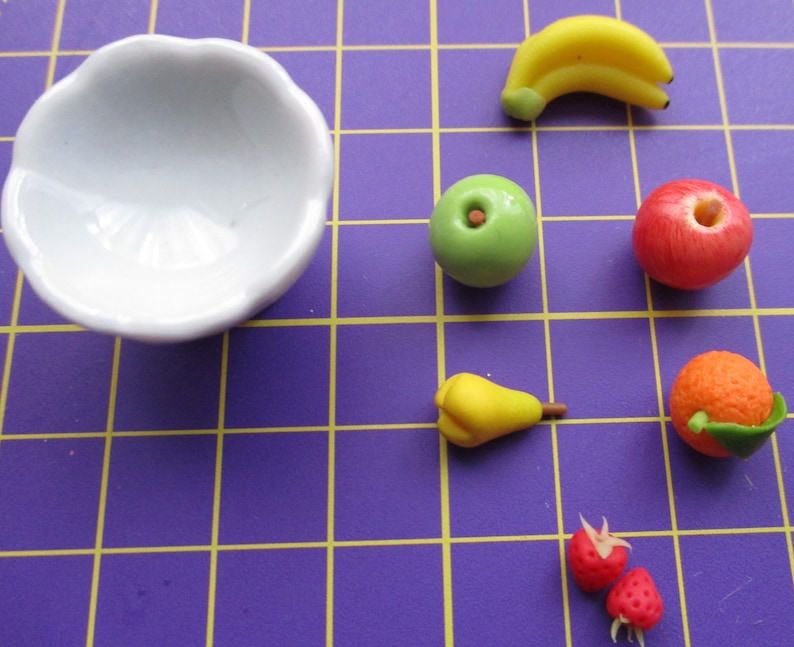 May include: A white, scalloped-edge bowl next to miniature fruit figurines. The fruit includes a yellow banana, green apple, red apple, yellow pear, orange, and two red strawberries. The fruit is arranged on a purple grid.