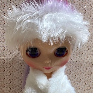 May include: A doll with a large head, prominent dark purple eyes, and long dark eyelashes. The doll wears a fluffy white faux fur hat that reveals purple hair underneath and a matching white faux fur collar around the neck, featuring pink blush.
