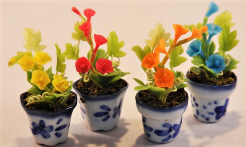 May include: Four miniature potted plants with colorful flowers in shades of yellow, red, orange, and blue. The small ceramic pots are white with blue floral patterns. The plants are set against a white background.