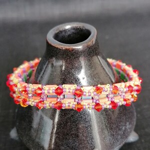 Könnte beinhalten: Ein mehrfarbiges Perlenarmband mit roten, orangefarbenen, gelben und violetten Perlen. Das Armband ist um eine kleine, dunkelbraune Keramikvase gewickelt.
